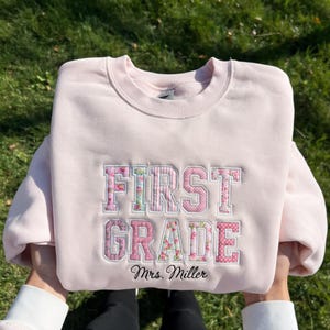 Puede incluir: Una sudadera de cuello redondo rosa claro con el texto FIRST GRADE bordado en letras grandes. Las letras están rellenas con patrones de flores y lunares rosas. Debajo del texto principal, el nombre Mrs. Miller está bordado en color negro.