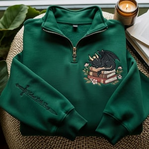 May include: A forest green quarter-zip sweatshirt featuring an embroidered design of a black dragon sleeping on a stack of books surrounded by flowers. The sleeve displays an embroidered sword with the text "Violent Little Thing" in black.