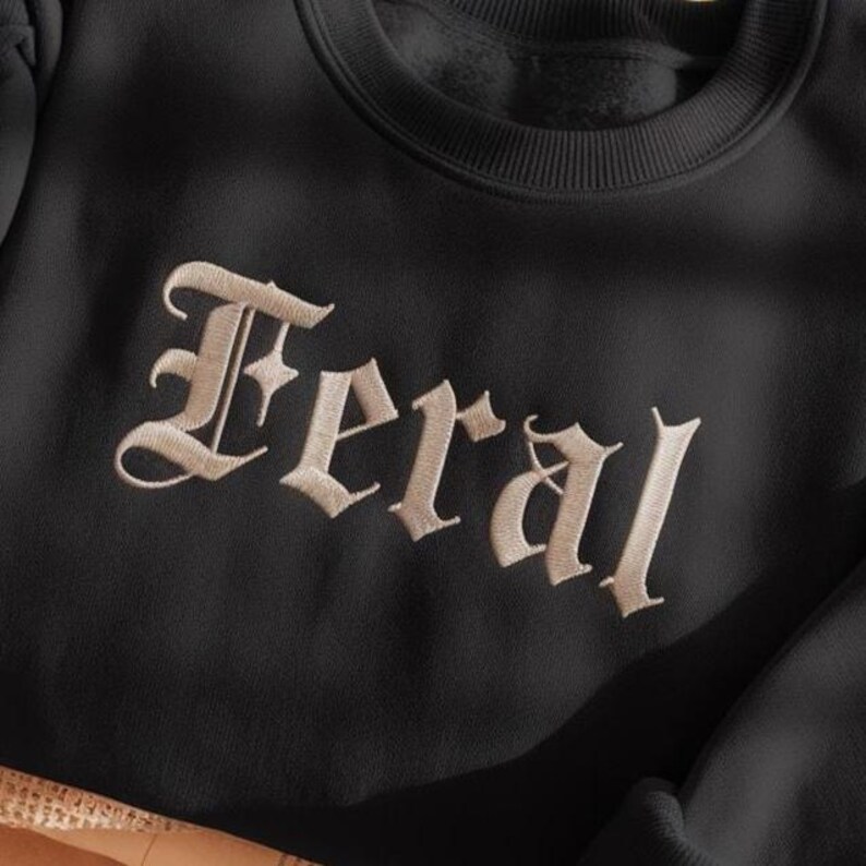 May include: Black sweatshirt with the word "Feral" embroidered in a light tan, gothic-style font. The text is centered on the chest of the garment. The sweatshirt has a crew neck and long sleeves.