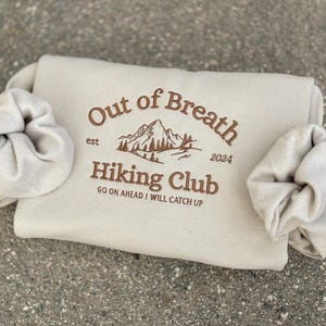 May include: A beige sweatshirt featuring brown embroidery that reads Out of Breath Hiking Club est 2024 with a mountain graphic and the text GO ON AHEAD I WILL CATCH UP. Two matching beige scrunchies are wrapped around the sides of the sweatshirt.