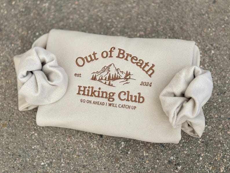 May include: A beige sweatshirt featuring brown embroidery that reads Out of Breath Hiking Club est 2024 with a mountain graphic and the text GO ON AHEAD I WILL CATCH UP. Two matching beige scrunchies are wrapped around the sides of the sweatshirt.