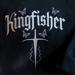 May include: Black sweatshirt with white embroidered design. The design features the word "Kingfisher" in a gothic font, a sword, and four butterflies. The sweatshirt has a crew neck and long sleeves.