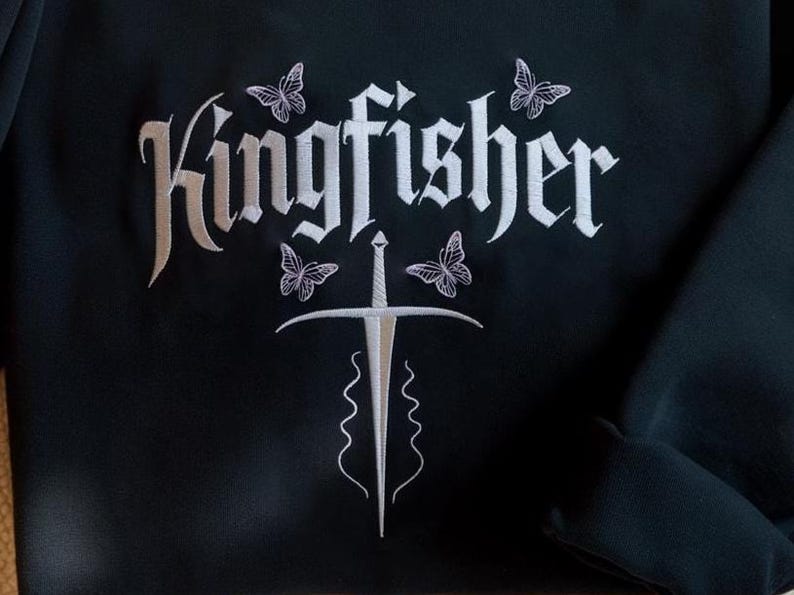 May include: Black sweatshirt with white embroidered design. The design features the word "Kingfisher" in a gothic font, a sword, and four butterflies. The sweatshirt has a crew neck and long sleeves.