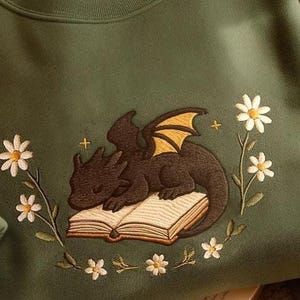 May include: A green sweatshirt with an embroidered design of a black dragon sleeping on an open book surrounded by white daisies.