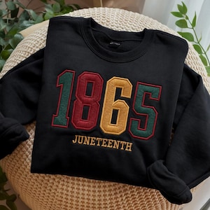 May include: Black sweatshirt with the embroidered text "1865" in red, gold, and green, with "JUNETEENTH" embroidered below. The sweatshirt is folded on a beige, textured surface.