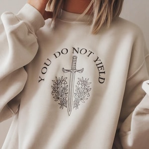 May include: A cream coloured sweatshirt with the text "You Do Not Yield" in a circle around a sword and floral design.