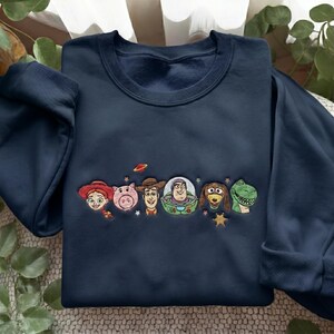 May include: A navy blue crewneck sweatshirt featuring an embroidered design of six animated characters from Toy Story, including a cowgirl, a pig, a cowboy, a space ranger, a dachshund, and a green dinosaur, accented with small stars and a planet.