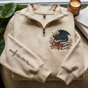 May include: A beige quarter-zip sweatshirt featuring an embroidered design of a black dragon with gold horns sleeping on a stack of books surrounded by flowers. The sleeve features an embroidered sword with the text Violent Little Thing.