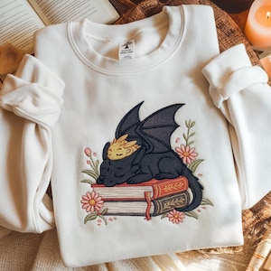 Puede incluir: Sudadera blanca con un diseño bordado de un dragón negro durmiendo sobre dos libros, rodeado de flores rosas y hojas verdes. El dragón tiene detalles dorados. La sudadera tiene mangas largas.