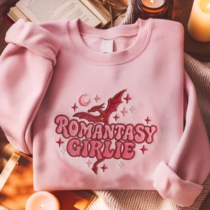 Könnte beinhalten: Hellrosa Sweatshirt mit dem gestickten Text "Romantasy Girlie" in Retro-Schrift, zusammen mit einem roten Drachen, Mond und Sternen. Das langärmlige Oberteil ist aus einem weichen Material gefertigt.