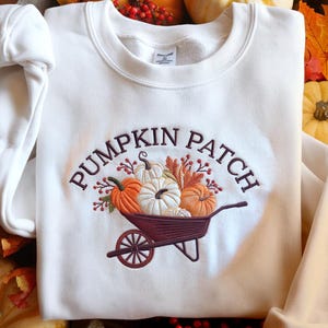 Może przedstawiać: Kremowa bluza z haftowanym wzorem. Wzór przedstawia napis "PUMPKIN PATCH" nad taczką wypełnioną dyniami i jesiennymi liśćmi. Dynie są pomarańczowe i białe.