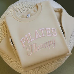 May include: A beige sweatshirt with the words "PILATES Therapy" embroidered in pink. The sweatshirt is folded and resting on a cream-colored, textured surface. The crew neck sweatshirt has long sleeves.