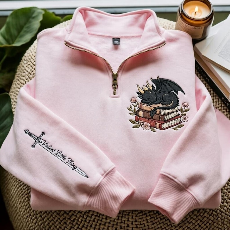 May include: A light pink quarter-zip sweatshirt featuring an embroidered black dragon sleeping on a stack of books with flowers. The right sleeve displays an embroidered silver sword with the text Violent Little Thing written along the blade.