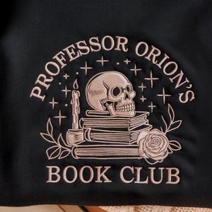 May include: Black fabric with an embroidered design featuring a skull, books, a candle, and a rose. The text reads "PROFESSOR ORION'S BOOK CLUB" in a light pink color, with stars and floral accents.