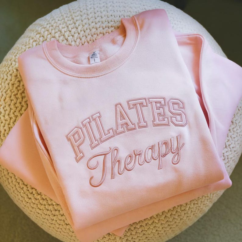 May include: A light pink sweatshirt with the words "PILATES Therapy" embroidered in a darker pink. The sweatshirt is folded and resting on a cream-colored, textured surface. The overall aesthetic is soft and feminine.