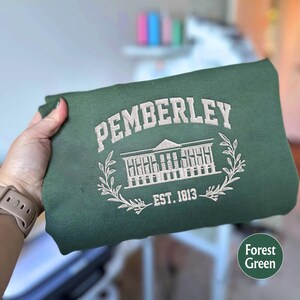 May include: A forest green sweatshirt with white embroidery. The embroidery features the text "Pemberley" in a cursive font, a building with windows, and the text "Est. 1813".