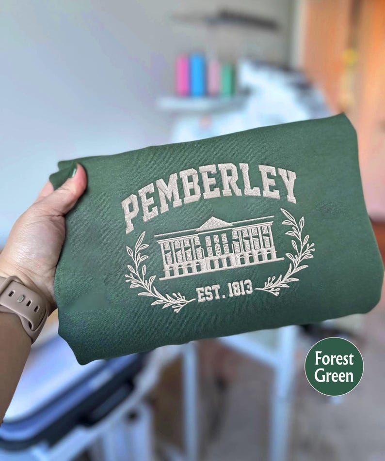 May include: A forest green sweatshirt with white embroidery. The embroidery features the text "Pemberley" in a cursive font, a building with windows, and the text "Est. 1813".