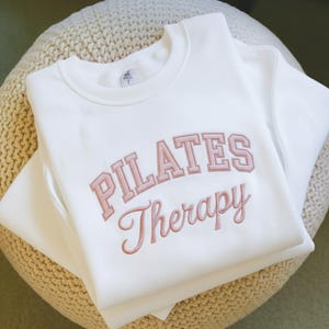 May include: A white crewneck sweatshirt with the words "PILATES Therapy" embroidered in a light pink color. The sweatshirt is folded and resting on a beige, textured surface.
