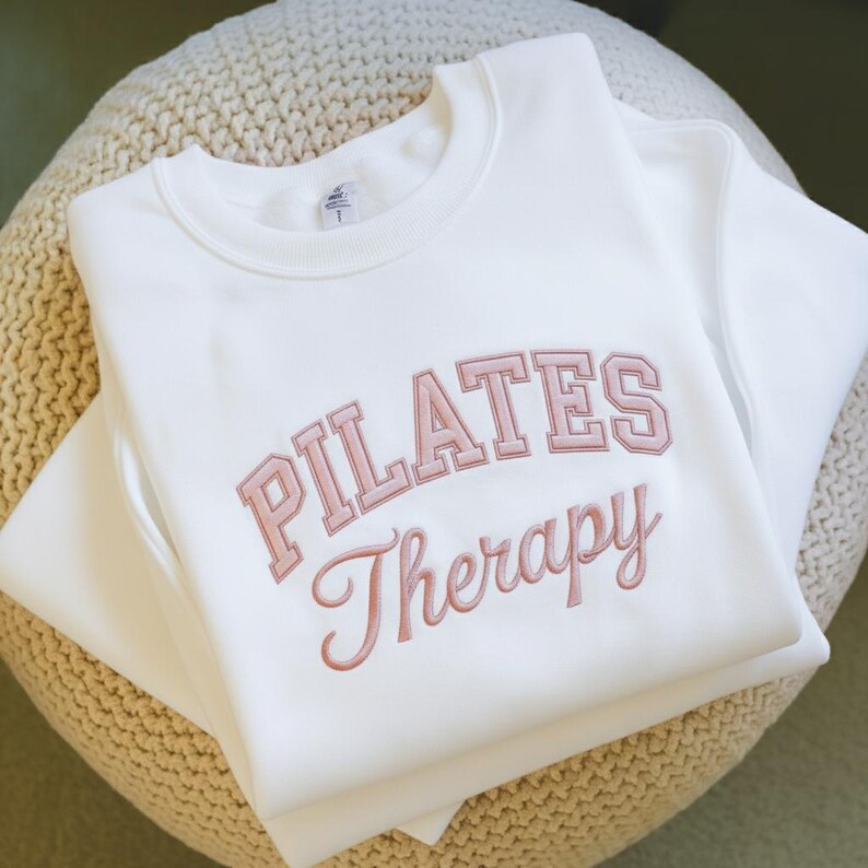 May include: A white crewneck sweatshirt with the words "PILATES Therapy" embroidered in a light pink color. The sweatshirt is folded and resting on a beige, textured surface.
