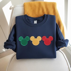 Puede incluir: Sudadera azul marino con tres dise&ntilde;os de cabeza de Mickey Mouse en verde, amarillo y rojo. La sudadera est&aacute; doblada sobre una silla con una manta amarilla en el fondo. El dise&ntilde;o es bordado.