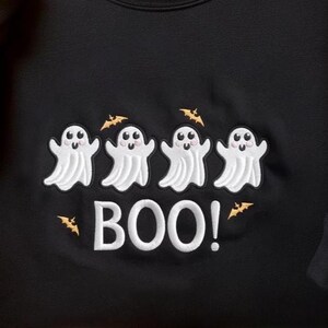 May include: A black sweatshirt featuring an embroidered design of four white ghosts with pink cheeks. Above the ghosts are small orange bats, and below them is the white embroidered text "BOO!" with an additional small orange bat to the right.