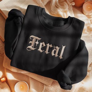 May include: Black crewneck sweatshirt with the word "Feral" embroidered in an ornate, off-white font. The sweatshirt is folded on a neutral-toned surface, with a book and a lit candle in the background.