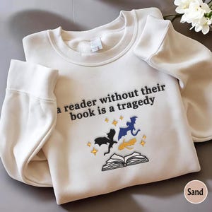 May include: A white sweatshirt with the text "A reader without their book is a tragedy" and an embroidered design of a book and three dragons.