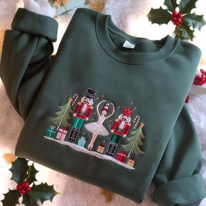 Puede incluir: Sudadera verde oscuro con un diseño navideño bordado. El diseño incluye dos cascanueces, una bailarina, árboles de Navidad, regalos y bastones de caramelo. La sudadera tiene mangas largas y cuello redondo.