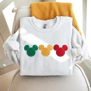 Puede incluir: Sudadera blanca de cuello redondo con tres dise&ntilde;os bordados de cabeza de rat&oacute;n en verde, amarillo y rojo. La sudadera est&aacute; doblada sobre una silla con una manta amarilla. La tela parece suave.