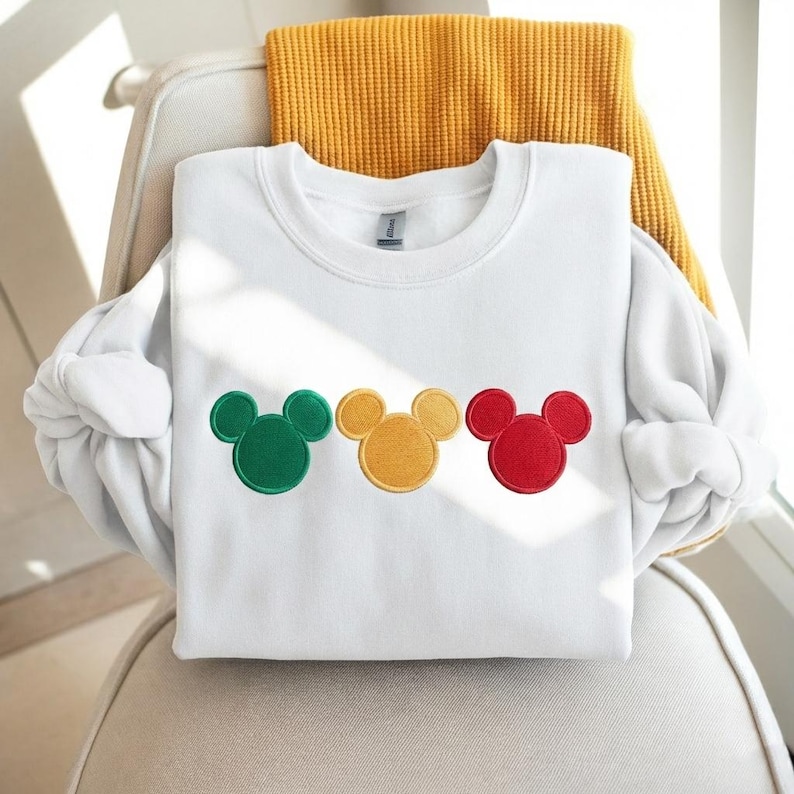 Puede incluir: Sudadera blanca de cuello redondo con tres dise&ntilde;os bordados de cabeza de rat&oacute;n en verde, amarillo y rojo. La sudadera est&aacute; doblada sobre una silla con una manta amarilla. La tela parece suave.