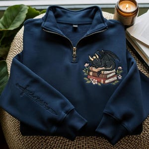 May include: A navy blue quarter-zip sweatshirt featuring an embroidered design of a black dragon sleeping on a stack of books surrounded by flowers. The sleeve displays an embroidered sword with the text "Velaris Little Thing" in script.
