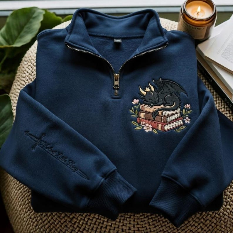 May include: A navy blue quarter-zip sweatshirt featuring an embroidered design of a black dragon sleeping on a stack of books surrounded by flowers. The sleeve displays an embroidered sword with the text "Velaris Little Thing" in script.