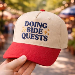 May include: A beige baseball cap with a red brim and a red button on top. The cap features the embroidered text "DOING SIDE QUESTS" in navy blue with gold star accents. The cap is held by a hand.