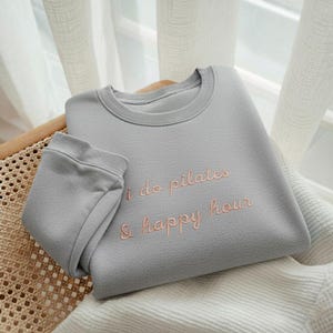 May include: A gray sweatshirt with the embroidered text "i do pilates & happy hour" in a light pink color. The sweatshirt is folded on a wicker chair.