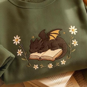 May include: A green sweatshirt with an embroidered design of a black dragon sleeping on an open book. The dragon has yellow wings and the book is surrounded by white daisies.