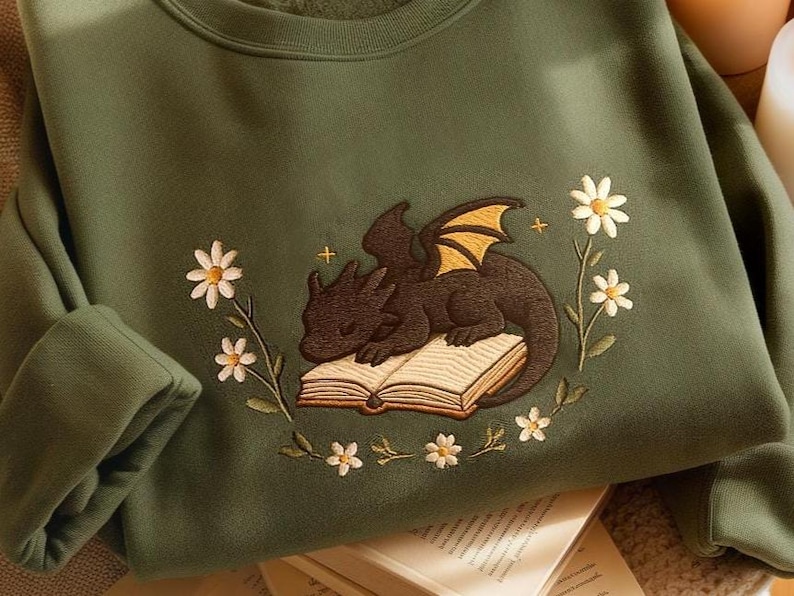 May include: A green sweatshirt with an embroidered design of a black dragon sleeping on an open book. The dragon has yellow wings and the book is surrounded by white daisies.