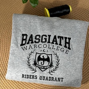 Könnte beinhalten: Grauer Sweatshirt mit einem schwarzen gestickten Design, das "Basgiath War College" mit einem Drachenlogo und den Worten "Fourth Wing Riders Quadrant" lautet.