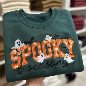 May include: A dark green crewneck sweatshirt featuring the embroidered text SPOOKY in orange and white checkered block letters, with the word season in black cursive script. White ghost figures, black bats, and spider webs are embroidered.