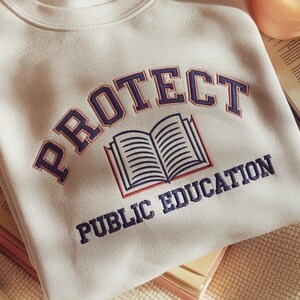 May include: A cream-coloured crewneck sweatshirt featuring dark blue embroidered text that reads PROTECT PUBLIC EDUCATION in an arched design, with a central graphic of an open book outlined in dark blue and red thread.