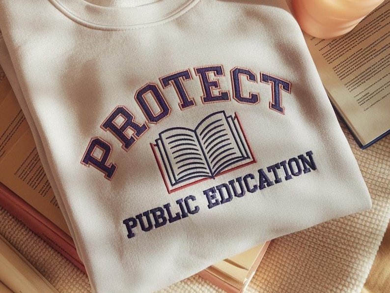 May include: A cream-coloured crewneck sweatshirt featuring dark blue embroidered text that reads PROTECT PUBLIC EDUCATION in an arched design, with a central graphic of an open book outlined in dark blue and red thread.