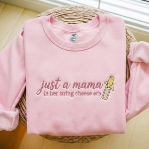 Könnte beinhalten: Hellrosa Sweatshirt mit der gestickten Aufschrift "just a mama in her string cheese era" und einer Käsesnack-Grafik. Das Sweatshirt ist aus weichem Material und hat einen Rundhalsausschnitt.