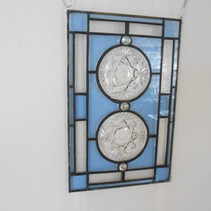 May include: A rectangular stained glass panel featuring light blue glass panes and two circular clear pressed glass inserts with a starburst pattern. The glass pieces are joined by dark metal lead lines, creating a geometric grid design.