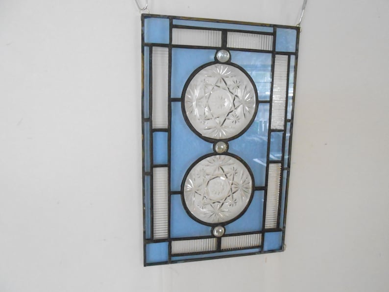 May include: A rectangular stained glass panel featuring light blue glass panes and two circular clear pressed glass inserts with a starburst pattern. The glass pieces are joined by dark metal lead lines, creating a geometric grid design.