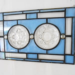 May include: A rectangular stained glass panel featuring a light blue border and center with two circular clear pressed glass inserts. The panel is framed in dark metal cames and includes three small clear glass cabochons along the horizontal center.