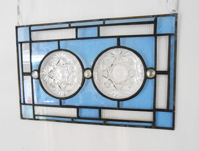May include: A rectangular stained glass panel featuring a light blue border and center with two circular clear pressed glass inserts. The panel is framed in dark metal cames and includes three small clear glass cabochons along the horizontal center.