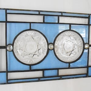 May include: A rectangular stained glass panel featuring a geometric frame of light blue and clear textured glass. Two circular, clear, pressed glass medallions with starburst patterns are centered within the frame, accented by small glass cabochons.