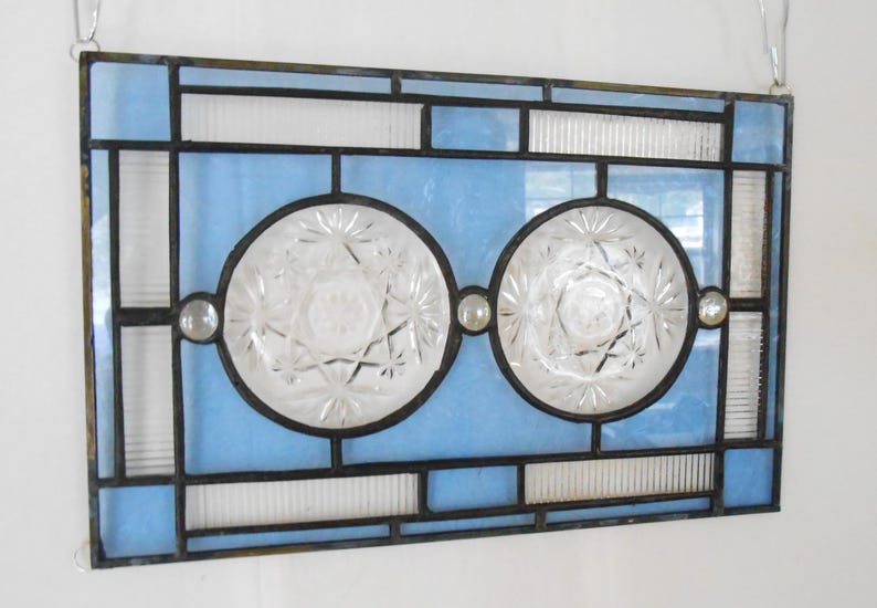 May include: A rectangular stained glass panel featuring a geometric frame of light blue and clear textured glass. Two circular, clear, pressed glass medallions with starburst patterns are centered within the frame, accented by small glass cabochons.