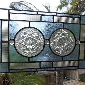May include: A rectangular stained glass panel featuring two central clear glass circles with starburst patterns, surrounded by textured light blue glass panes and vertical ribbed glass accents, all held together by a dark metal lead came frame.