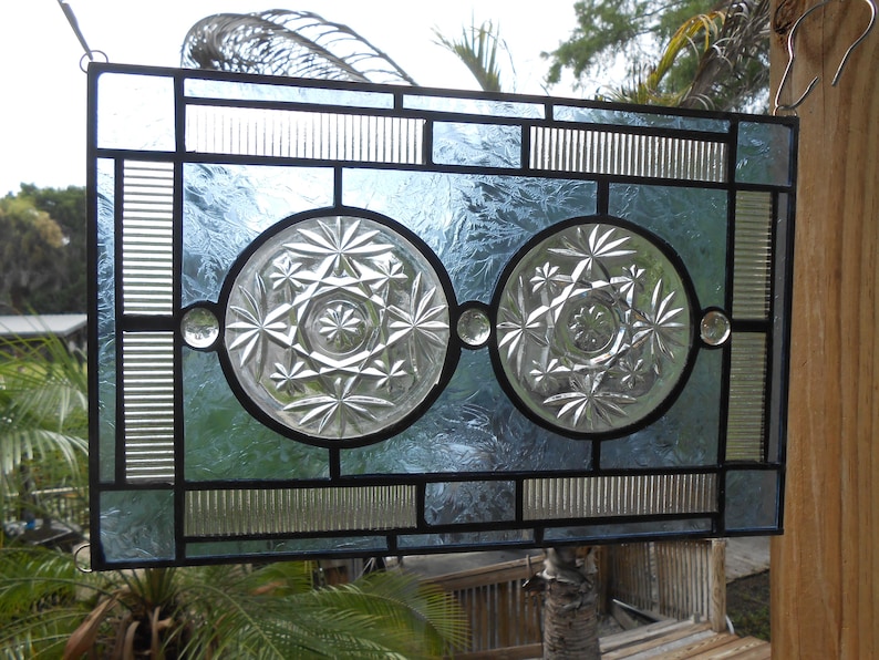 May include: A rectangular stained glass panel featuring two central clear glass circles with starburst patterns, surrounded by textured light blue glass panes and vertical ribbed glass accents, all held together by a dark metal lead came frame.