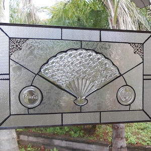 May include: A stained glass window featuring a clear glass fan design with a black metal frame. The fan is made of clear glass with a textured surface. The window is hanging from a hook.